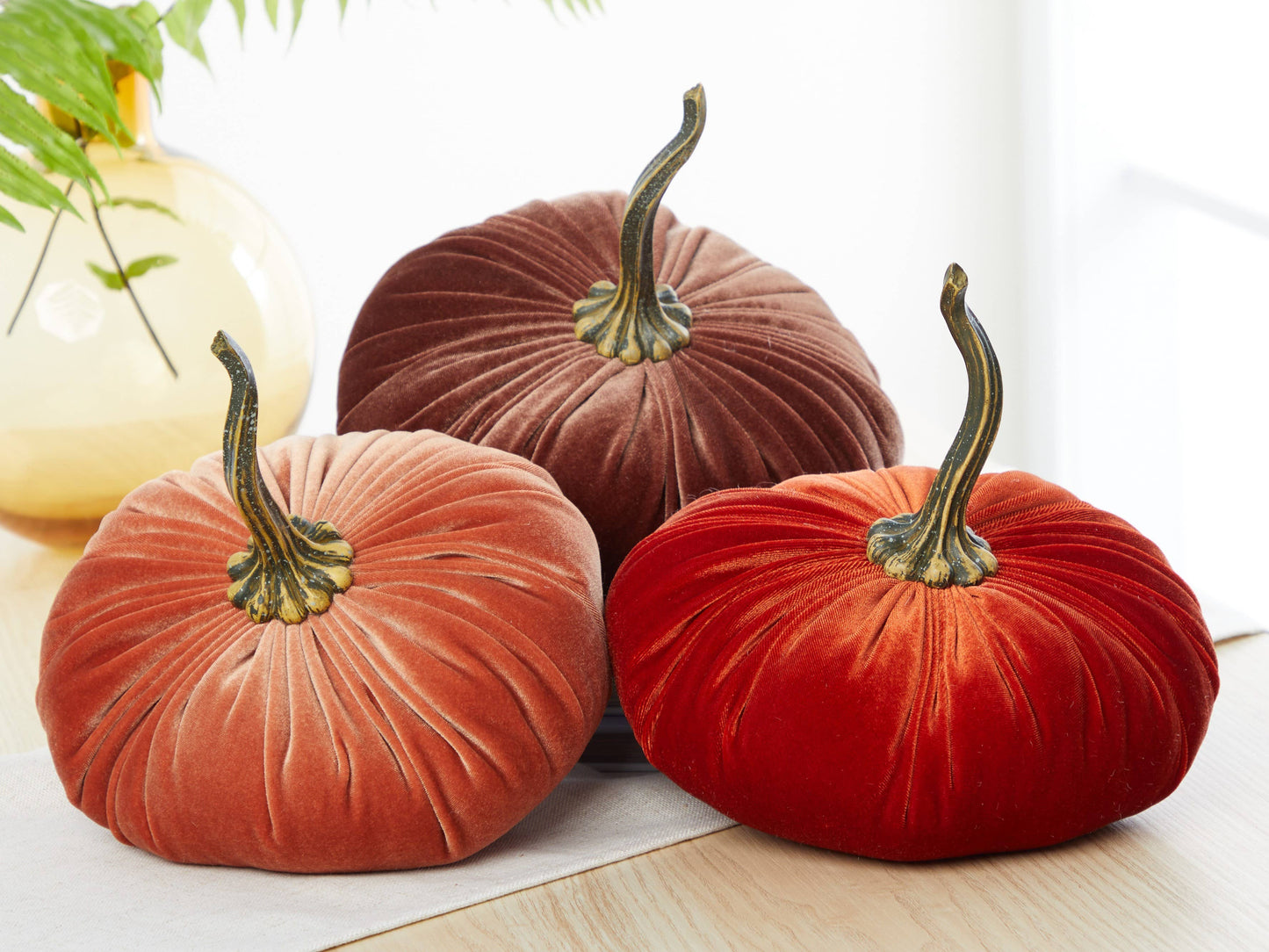 Three extra-large velvet pumpkins from Your Heart’s Content, in rich shades of orange, brown, and red with realistic curved stems, are displayed on a light wooden surface beside a vase and green leaves—making them an elegant fall accent or thoughtful gift for weddings and holiday mantel décor.