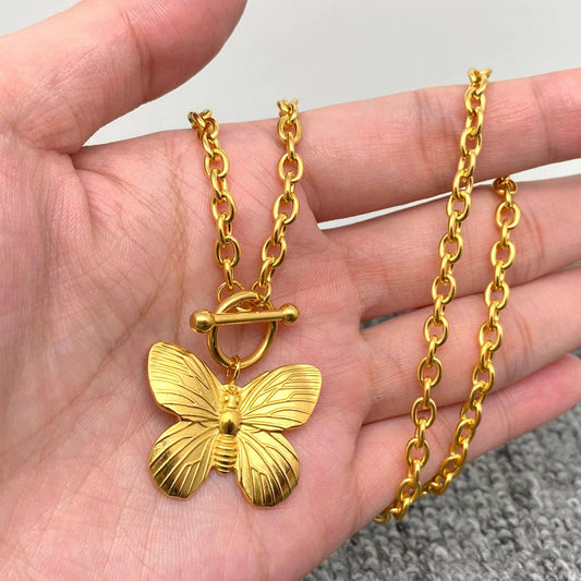 A hand holds the Mio Queena - Butterfly 18K Gold Plated Steel Buckle Necklace by Mio Queena, featuring a detailed gold butterfly pendant and a stylish toggle clasp. The background is simple and neutral.