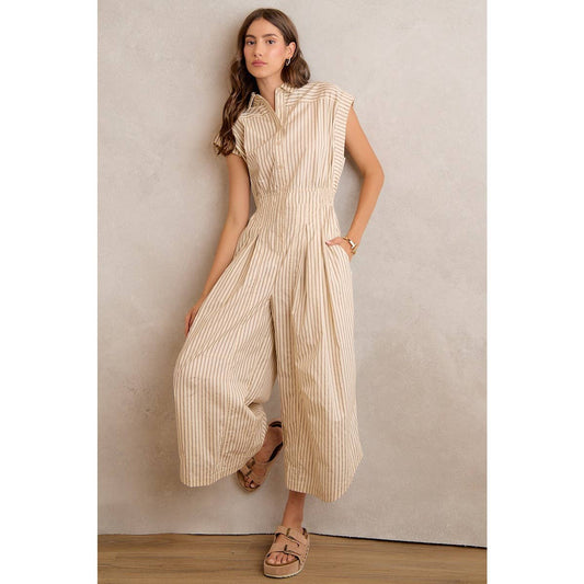 A woman stands against a neutral wall with one hand in her pocket, wearing the So Me - SLEEVELESS WIDE LEG STRIPE JUMPSUIT by So Me in beige and white stripes, paired with sandals.