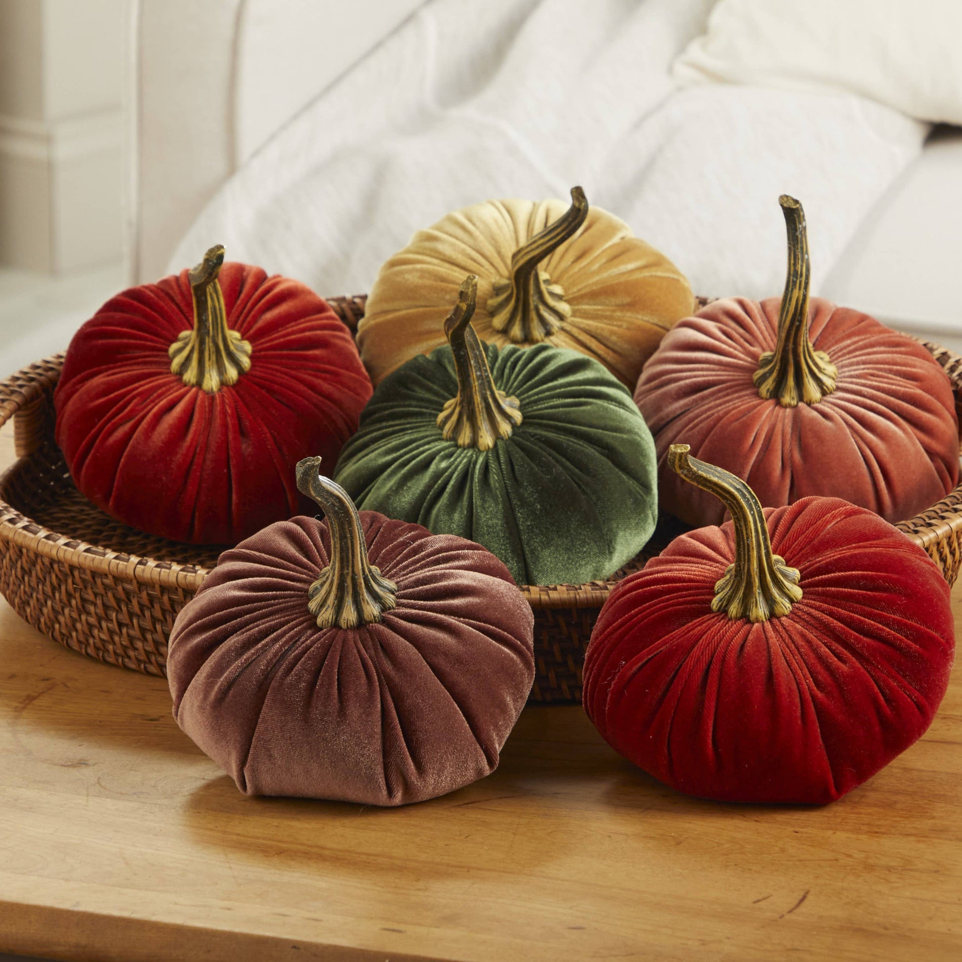 Six large velvet pumpkins from Your Heart’s Content, in assorted fall colors like red, green, gold, orange, and brown, are beautifully arranged in a woven basket on a wooden table with a white sofa in the background—creating an eye-catching handcrafted decor piece with Your Heart’s Content Pumpkin Large Velvet that’s perfect as a Thanksgiving or Halloween centerpiece.