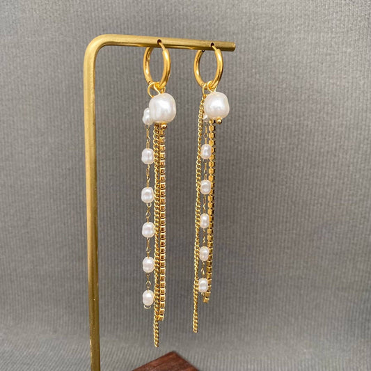 Introducing the Mio Queena - Zircon & Pearl Fringe 18K Gold Plated SS Huggie Earrings-FGS by Mio Queena. These elegant earrings feature gold hoops adorned with dangling gold chains and delicate white pearls, beautifully displayed on a gold stand against a gray textured background. Crafted from stainless steel and finished with 18K gold plating, they are designed to be hypoallergenic—making them a perfect choice for sensitive ears.