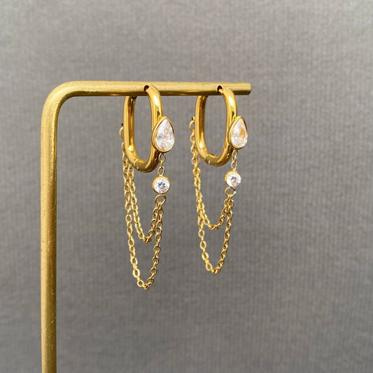 Here’s a rewritten version using your product data: The Mio Queena - Chain Fringe Zircon Inlaid 18K Gold Plated SS Earrings (FGS) feature gold hoops adorned with teardrop and round zircon crystals, each elegantly accented by two delicate dangling gold chains. These non-tarnish earrings by Mio Queena are displayed on a gold earring stand against a plain gray background, offering lasting shine and durability.