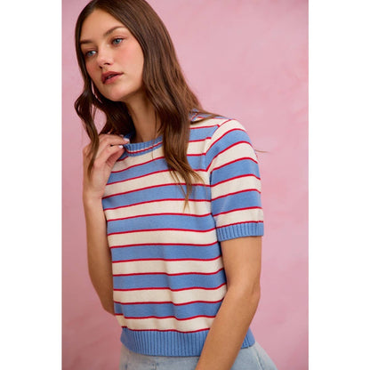 A woman with long brown hair stands against a pink background, looking slightly to the side with one hand near her neck. She is wearing the So Me STRIPE KNIT CREW NECK SHORT SLEEVES SWEATER TOP—a trendy women's piece by So Me featuring light blue and white stripes with red lines and a crew neckline.