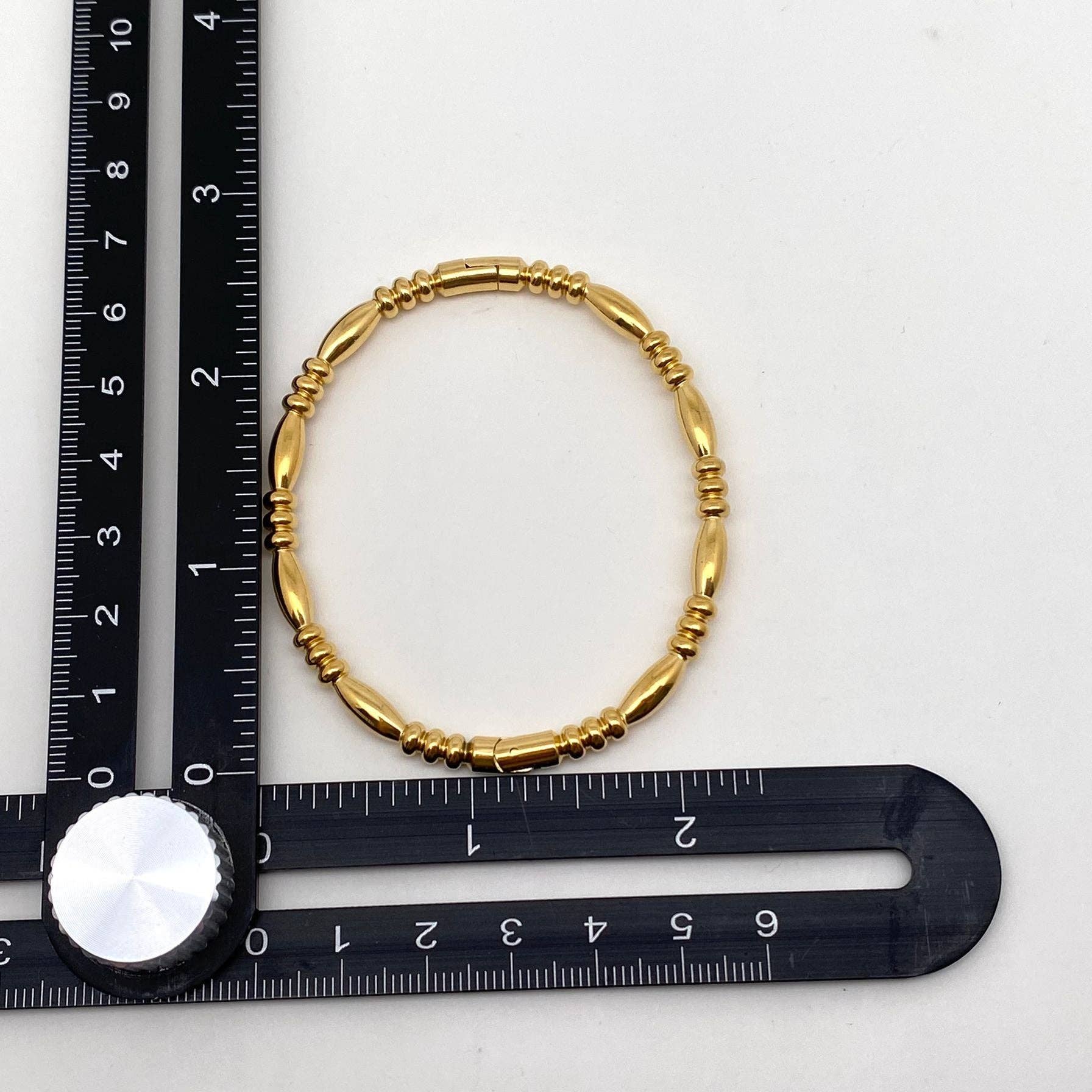 A Mio Queena Multi-style 18K Gold-plated Stainless Steel Bangle is positioned at the intersection of two black measuring rulers arranged at a right angle on a white surface.