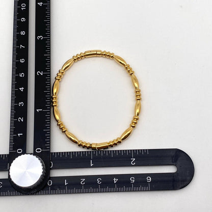 A Mio Queena Multi-style 18K Gold-plated Stainless Steel Bangle is positioned at the intersection of two black measuring rulers arranged at a right angle on a white surface.