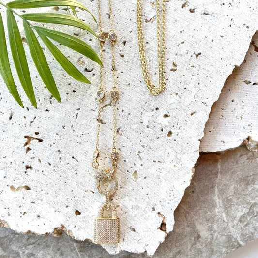 VB&CO Designs gold lock necklace styled on white stone with green foliage accent