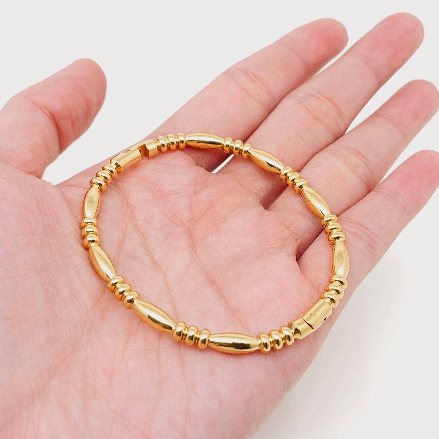 A hand holding the Mio Queena - Multi-style 18K Gold-plated Stainless Steel Bangle (FGS), featuring alternating smooth and ribbed oval beads, displayed against a white background—an elegant piece by Mio Queena perfect for jewelry gifting.