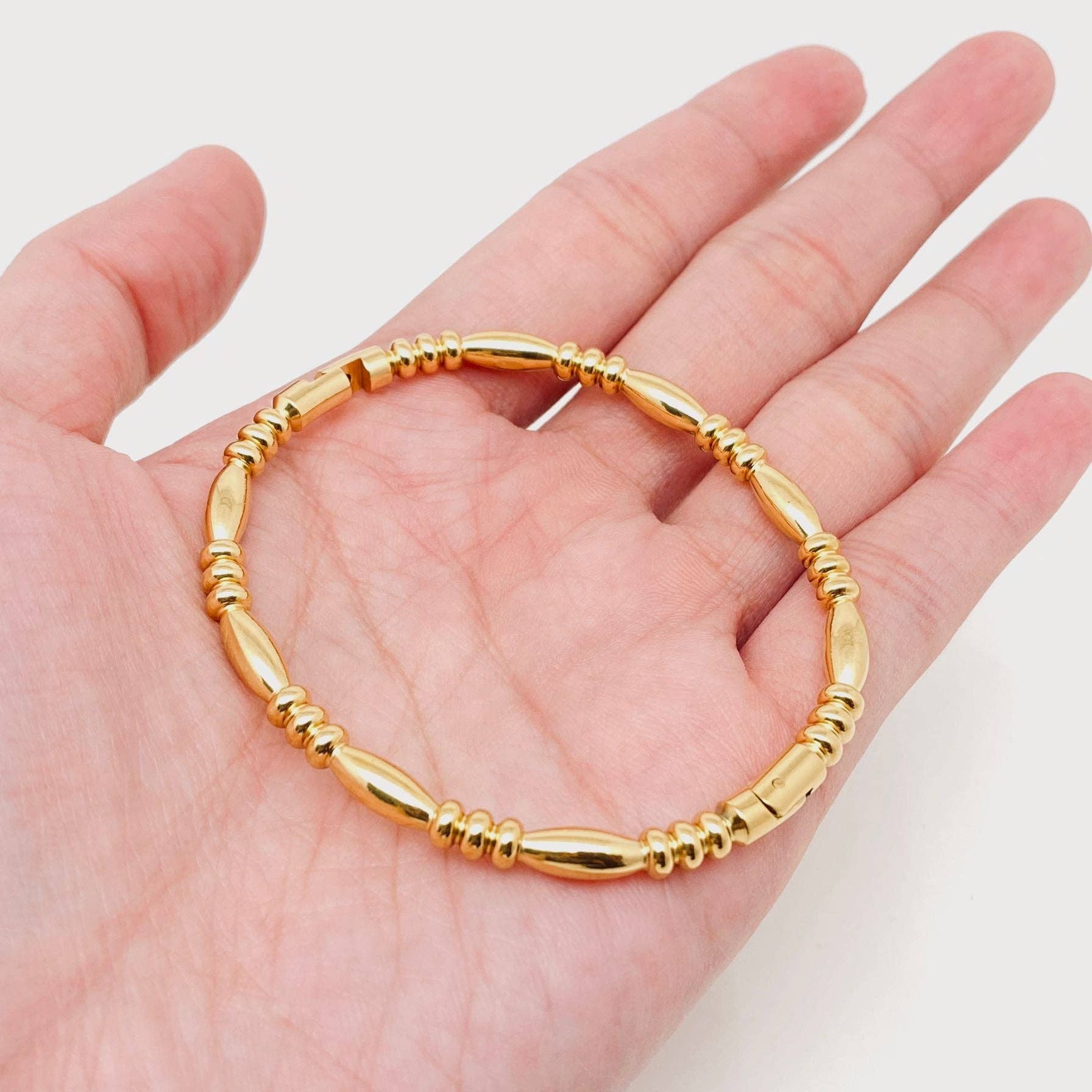 A hand holding the Mio Queena - Multi-style 18K Gold-plated Stainless Steel Bangle (FGS), featuring alternating smooth and ribbed oval beads, displayed against a white background—an elegant piece by Mio Queena perfect for jewelry gifting.