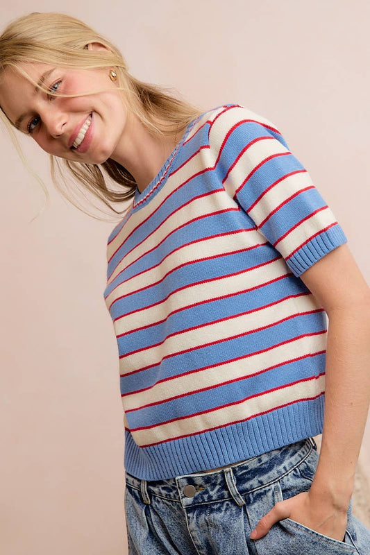 A smiling person with long blonde hair wears the So Me STRIPE KNIT CREW NECK SHORT SLEEVES SWEATER TOP, featuring blue, white, and red stripes, paired with high-waisted blue jeans, standing against a pale pink background.