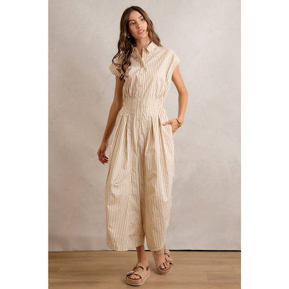 A woman stands on a wooden floor in a So Me SLEEVELESS WIDE LEG STRIPE JUMPSUIT featuring beige and white stripes, a collared neck, and wide legs. She has long wavy hair, wears platform sandals, and is set against a neutral background.