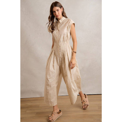 A woman models the So Me SLEEVELESS WIDE LEG STRIPE JUMPSUIT by So Me, featuring a beige hue, subtle stripe detailing, button accents, and wide legs. She pairs the look with tan sandals while standing on a wooden floor against a neutral backdrop.