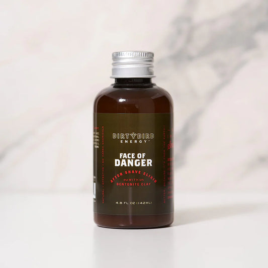 A brown bottle labeled "Dirty Bird Face of Danger After-Shave Elixir" by Dirty Bird, featuring Bentonite Clay and witch hazel, sits on a white surface against a light marble background, showcasing this natural aftershave solution.