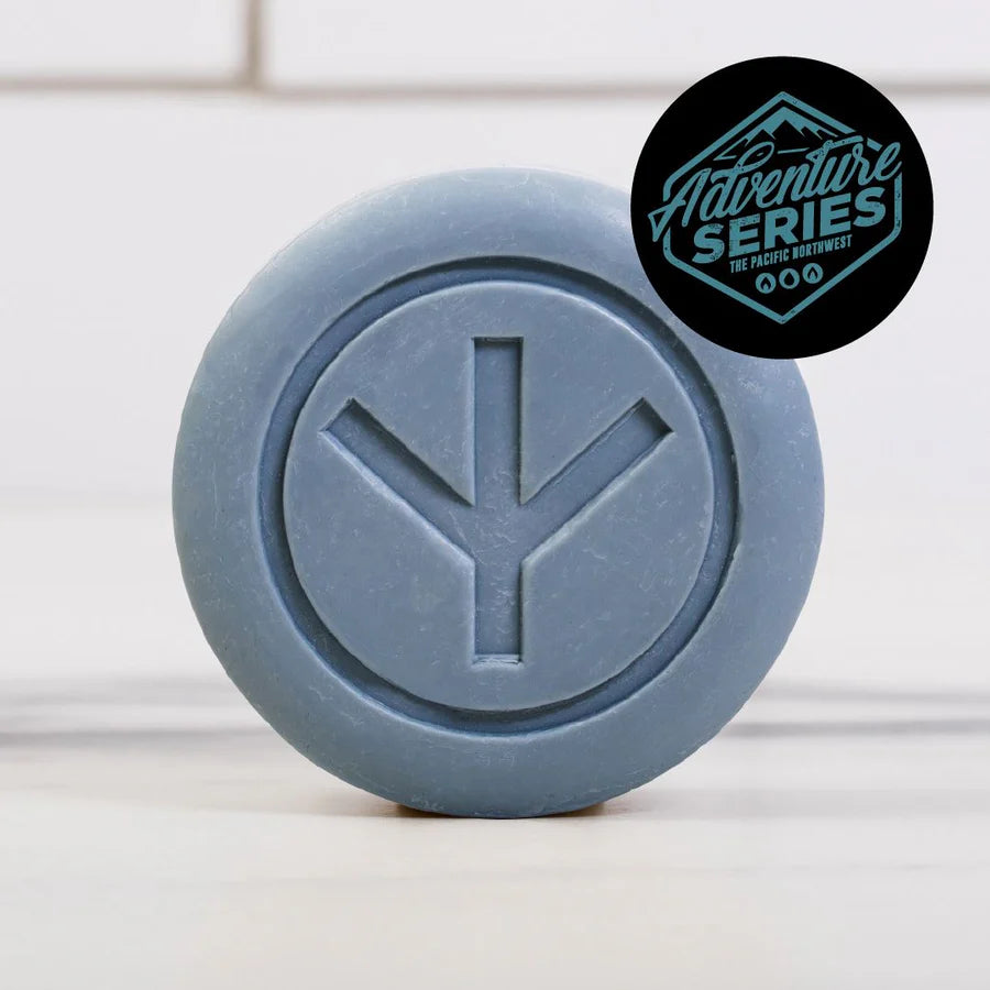 A round blue Dirty Bird Bar Soap from Dirty Bird, featuring a geometric symbol engraved on its surface, stands on a light background. Made with natural ingredients, this soap provides gentle cleansing. A black badge reading "Adventure Series The Pacific Northwest" appears in the corner.