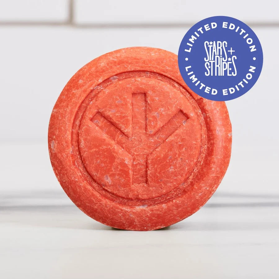 A round, red-orange Dirty Bird Bar Soap with a peace symbol embossed on top. Crafted as a natural soap by Dirty Bird, this recovery bar gently cleanses while promoting revitalization. A blue sticker in the corner reads “LIMITED EDITION STARS + STRIPES LIMITED EDITION.” The exfoliating bar rests on a light surface.