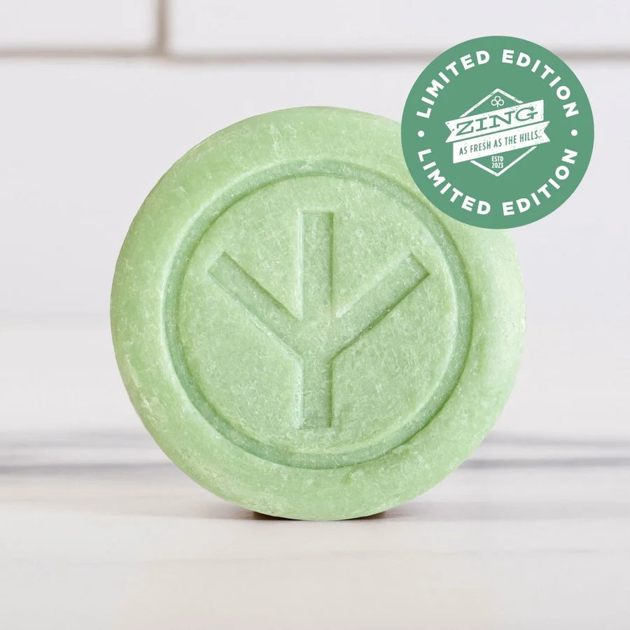 A round, green bar of Dirty Bird Bar Soap with a tree-like symbol embossed on it. A green sticker in the corner reads "Limited Edition Zing As Fresh As The Hills." The soap is set against a background of white tile.