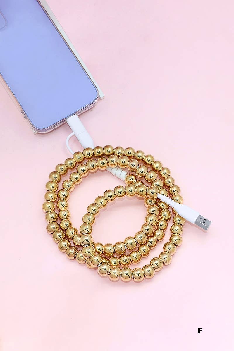 A phone with a lavender case is charging using the 42POPS Beaded Dual USB Fast Charging Cable by 42POPS, featuring shiny gold beads and coiled on a light pink surface.