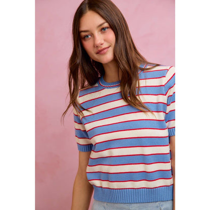 A woman with long brown hair stands in front of a pink background, wearing the So Me STRIPE KNIT CREW NECK SHORT SLEEVES SWEATER TOP from So Me—a trendy blue and white stripe knit sweater with red accents.
