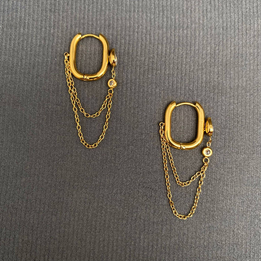 Mio Queena's Chain Fringe Zicon Inlaid 18K Gold Plated SS Earrings – FGS feature two non-tarnish hoop earrings, each embellished with double chain fringes and a small crystal charm. Displayed on a textured gray surface, these elegant and durable earrings offer everyday style combined with the lasting shine of 18K gold plating.