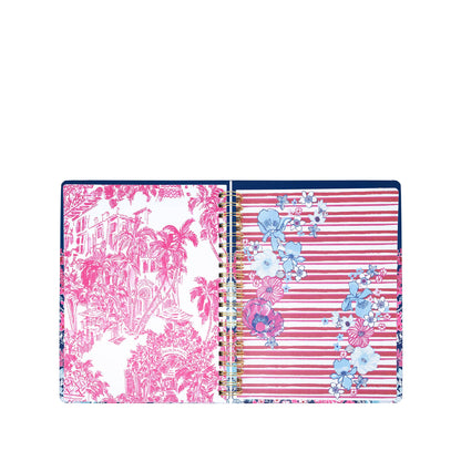 The Lilly Pulitzer by Lifeguard Press - Jumbo 17 Month Agenda, In All Her Glory, is an open spiral-bound planner displaying a page with pink tropical landscape art and another featuring pink stripes accented with blue, white, and pink floral designs.