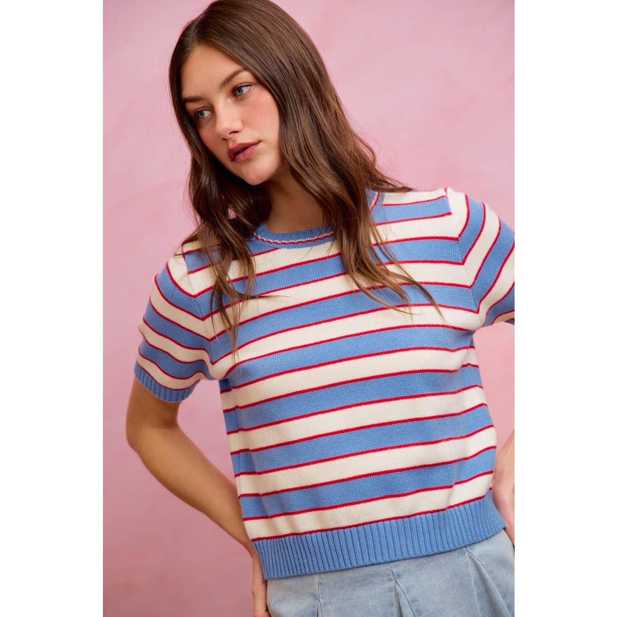 A woman with long brown hair stands in front of a pink background wearing the So Me - STRIPE KNIT CREW NECK SHORT SLEEVES SWEATER TOP by So Me, a trendy women’s sweater featuring light blue, red, and white horizontal stripes.