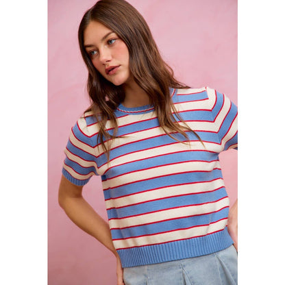 A woman with long brown hair stands in front of a pink background wearing the So Me - STRIPE KNIT CREW NECK SHORT SLEEVES SWEATER TOP by So Me, a trendy women’s sweater featuring light blue, red, and white horizontal stripes.