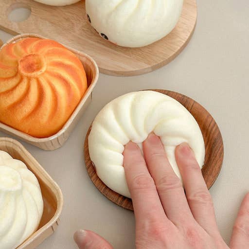 Hand pressing white K-Wonderland dumpling squishy fidget on wooden tray with orange grilled dumpling nearby