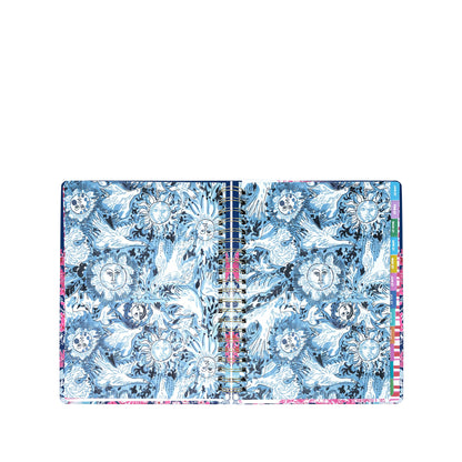 A Lilly Pulitzer by Lifeguard Press Jumbo 17 Month Agenda, In All Her Glory, lies open to reveal blue and white pages adorned with intricate sun and moon designs, while colored tab dividers from the planner are visible along the right edge.