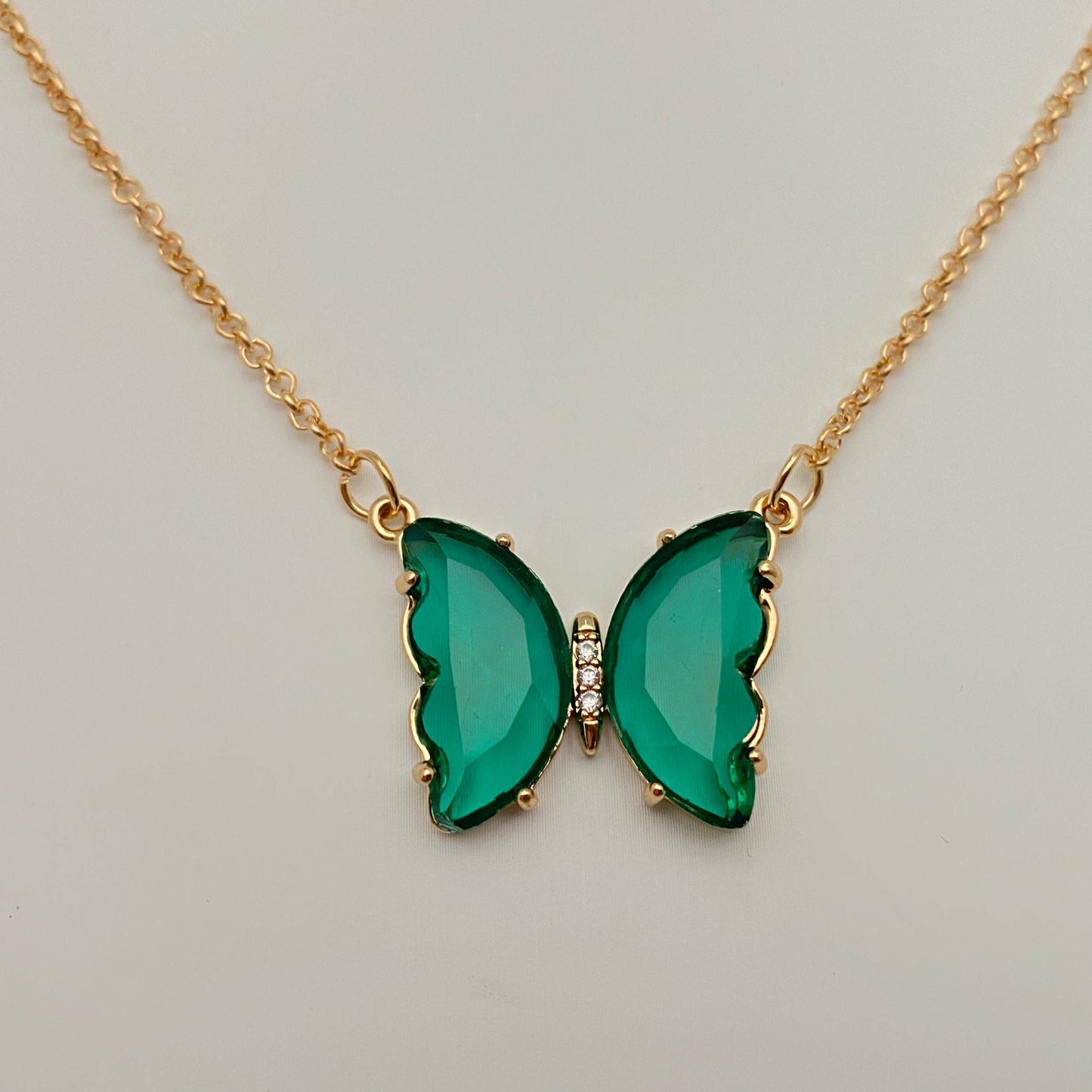 The Mio Queena - Multicolor Glass Butterfly Pendant Necklace by Mio Queena features delicate green gemstone wings and sparkling clear stones at its center. Displayed on a neutral background, this elegant necklace is crafted as hypoallergenic jewelry, making it perfect for sensitive skin.
