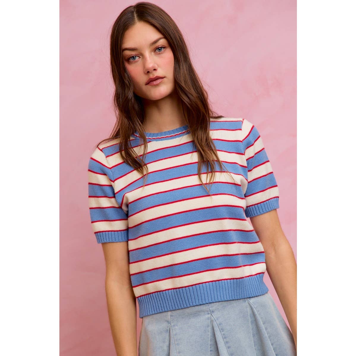 A woman with long brown hair wears the So Me STRIPE KNIT CREW NECK SHORT SLEEVES SWEATER TOP, featuring a trendy light blue fabric with bold red and white horizontal stripes and a classic crew neck short sleeve design. She pairs it with a blue skirt while standing against a pink background, showcasing the modern and stylish look of this So Me women's top.