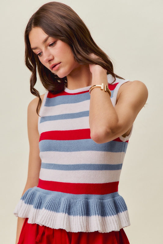 A woman with wavy brown hair wearing the So Me 4th Of July Stripe Ruffle Bottom Sweater Knit Top—a sleeveless knit top from So Me featuring red, white, and blue stripes with a blue ruffled hem—looks down and touches her hair against a neutral background. This festive top is perfect for patriotic parties or 4th of July celebrations.