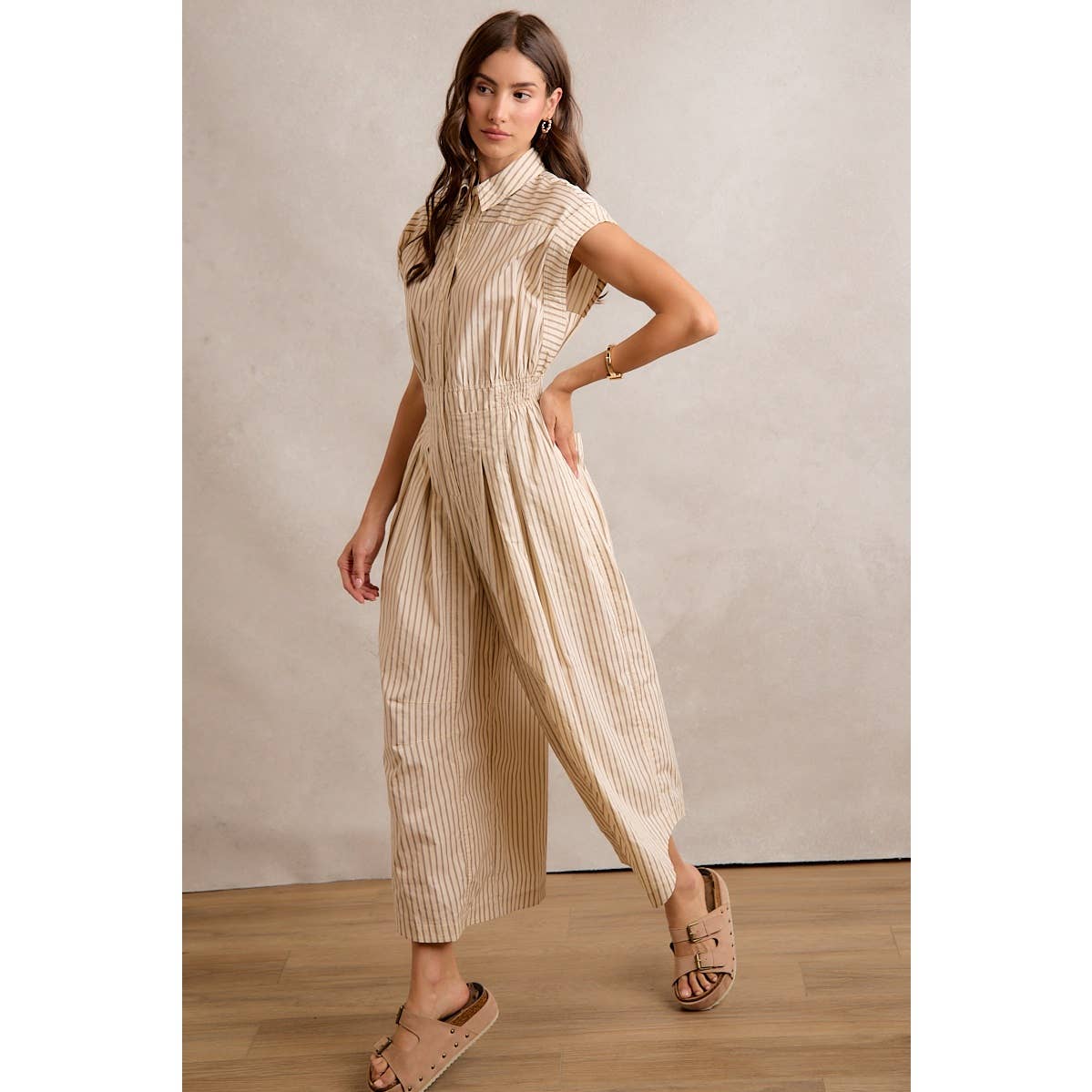 A woman poses indoors wearing the So Me Sleeveless Wide Leg Stripe Jumpsuit in beige and white, paired with tan slide sandals. She stands on a wooden floor against a neutral background.