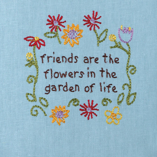 Close-up of embroidered flower design on light blue kitchen towel with red, yellow, and purple blooms surrounding friendship sentiment