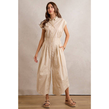 A woman stands on a wooden floor against a neutral textured background, hands in the pockets of the So Me SLEEVELESS WIDE LEG STRIPE JUMPSUIT by So Me—featuring beige and white stripes, wide cropped legs, and paired with tan sandals.