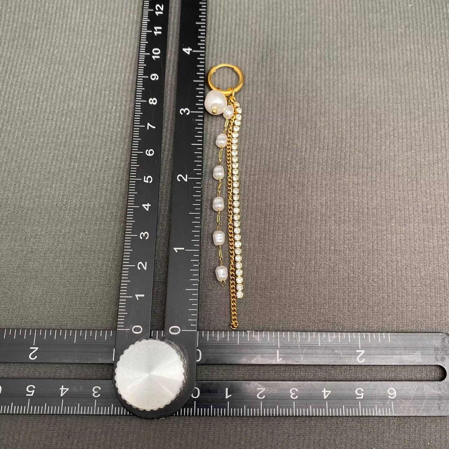 The Mio Queena - Zircon & Pearl Fringe 18K Gold Plated SS Huggie Earrings-FGS by Mio Queena are displayed on a T-square ruler, revealing the elegant earrings to be approximately 4 inches long against a textured grey background—a chic addition perfect for complementing your favorite hypoallergenic jewelry.