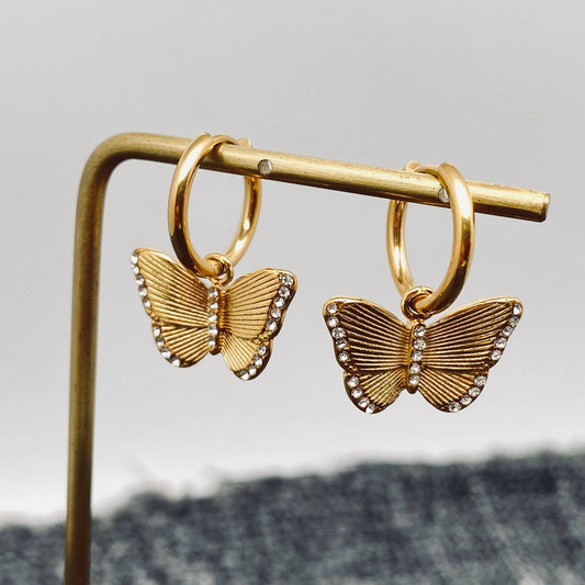 The Mio Queena - Butterfly Gold Plated Huggie Earring by Mio Queena features gold hoop earrings adorned with butterfly charms, each accented with sparkling clear rhinestones on the wings. Displayed on a gold stand against a softly blurred gray fabric background, these hypoallergenic earrings are crafted from stainless steel and finished with an 18K gold plating—offering both elegance and comfortable wear for sensitive ears.