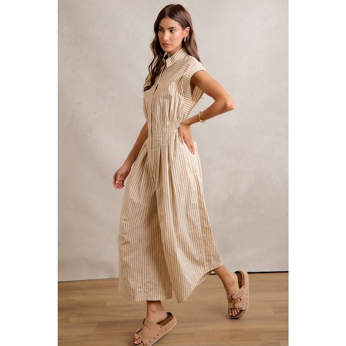 A woman stands sideways on a wooden floor against a light background wearing the So Me SLEEVELESS WIDE LEG STRIPE JUMPSUIT, featuring wide legs, pockets, and chic beige stripes. With one hand in her pocket and tan slide sandals completing the look, she showcases effortless style in this jumpsuit by So Me.