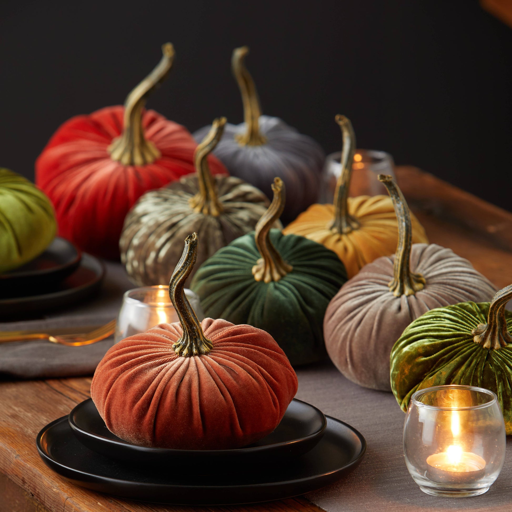 Small velvet pumpkins from Your Heart’s Content are arranged in a variety of colors on a wooden table, complemented by black plates, gray napkins, and glowing tealight candles to create a warm and cozy autumnal setting—making them the perfect centerpiece for your Thanksgiving or Valentine’s mantel.