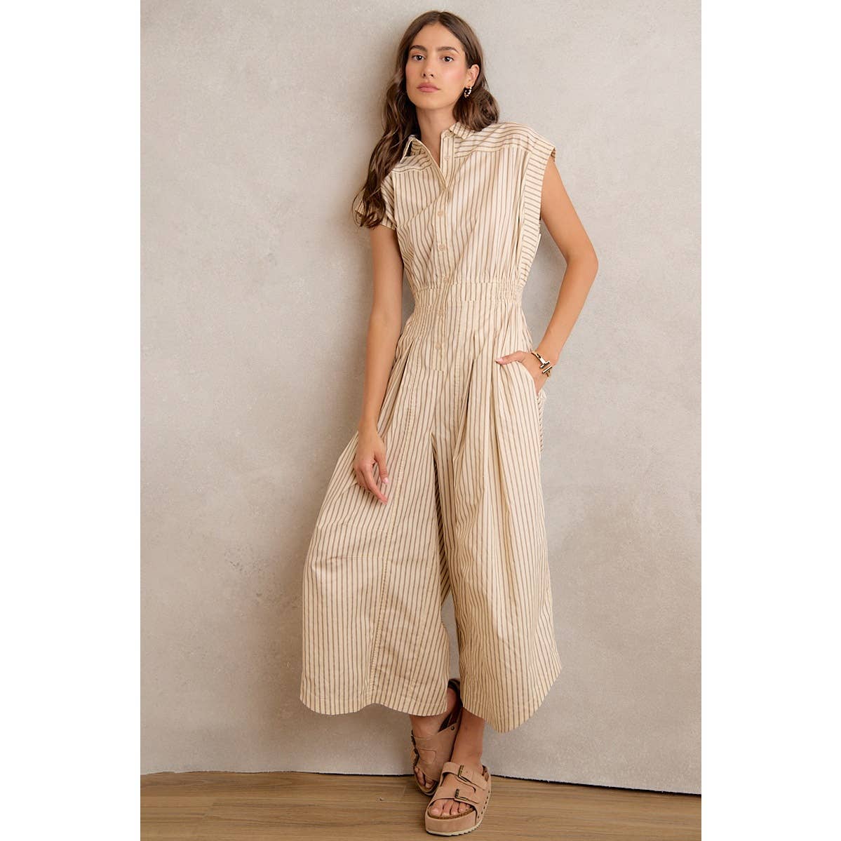 A woman stands against a beige wall, hands in her pockets, wearing the So Me SLEEVELESS WIDE LEG STRIPE JUMPSUIT with thin vertical stripes, styled with sandals and bracelets.