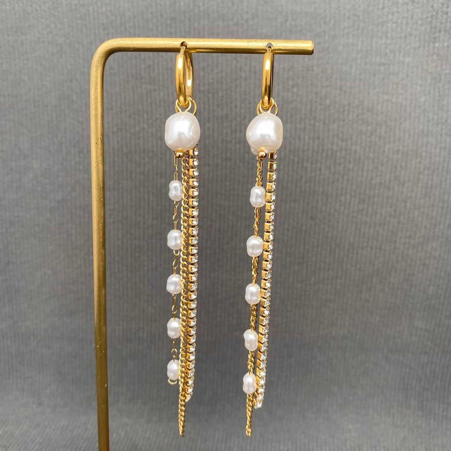 Introducing the Mio Queena - Zircon & Pearl Fringe 18K Gold Plated SS Huggie Earrings-FGS by Mio Queena. These hypoallergenic huggie earrings feature elegant gold hoops adorned with dangling strands of pearls and sparkling zircon stones, all crafted from durable 18K gold plated stainless steel for lasting shine. Displayed on a gold stand against a gray textured background, these stylish jewelry pieces effortlessly combine sophistication with everyday wearability.