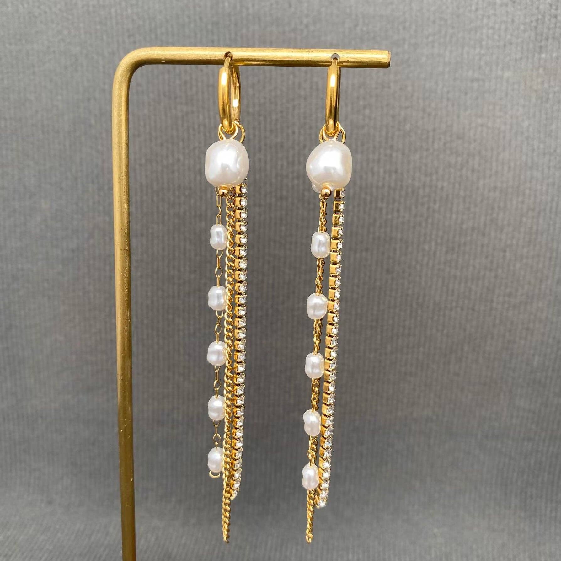 Introducing the Mio Queena - Zircon & Pearl Fringe 18K Gold Plated SS Huggie Earrings-FGS by Mio Queena. These hypoallergenic huggie earrings feature elegant gold hoops adorned with dangling strands of pearls and sparkling zircon stones, all crafted from durable 18K gold plated stainless steel for lasting shine. Displayed on a gold stand against a gray textured background, these stylish jewelry pieces effortlessly combine sophistication with everyday wearability.