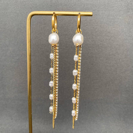 Introducing the Mio Queena - Zircon & Pearl Fringe 18K Gold Plated SS Huggie Earrings-FGS by Mio Queena. These hypoallergenic huggie earrings feature elegant gold hoops adorned with dangling strands of pearls and sparkling zircon stones, all crafted from durable 18K gold plated stainless steel for lasting shine. Displayed on a gold stand against a gray textured background, these stylish jewelry pieces effortlessly combine sophistication with everyday wearability.