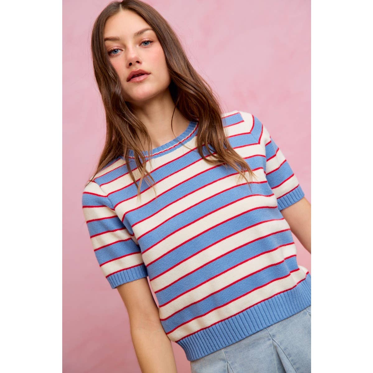 A woman with long brown hair is wearing the So Me STRIPE KNIT CREW NECK SHORT SLEEVES SWEATER TOP and light blue jeans, standing in front of a pink background.