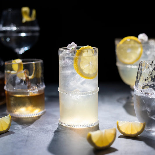 A Juliska "Graham" Highball Glass filled with a clear cocktail, ice, lemon slices, and garnished with a small white flower sits on a table, surrounded by other drinks and lemon wedges.