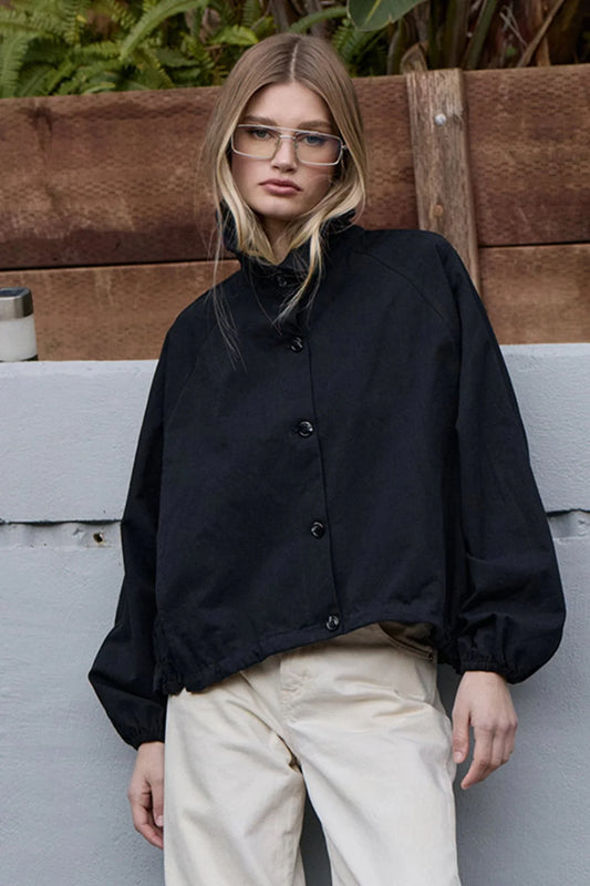 Raisonnel High Neck Button-up Jacket in black, worn with cream pants, front view