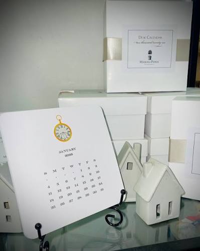 The Maison de Papier Desk Calendar 2026 by maison de papier is displayed on a stand beside miniature white house ornaments and stacked white boxes in the background.