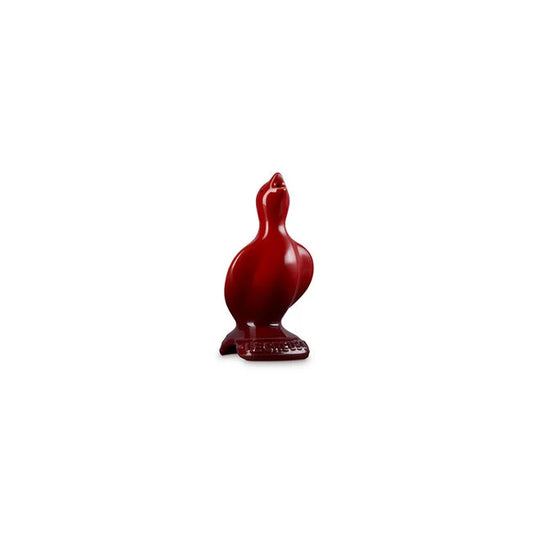 The Le Creuset Heritage Pie Bird Cerise by Le Creuset is a glossy red ceramic figurine that stands upright on a rectangular base with textured lettering, its beak pointed upward. The background is plain white.