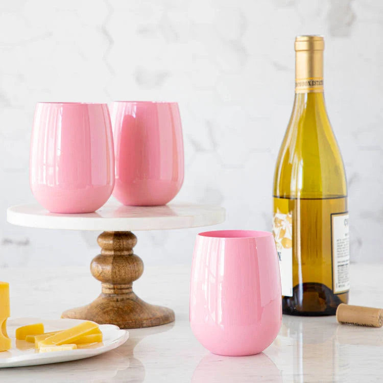 Three unbreakable SymGlass Stemless Wine Glasses in pink are displayed on a wooden cake stand beside a bottle of white wine, a cork, and a plate with sliced cheese, all arranged on a marble countertop.