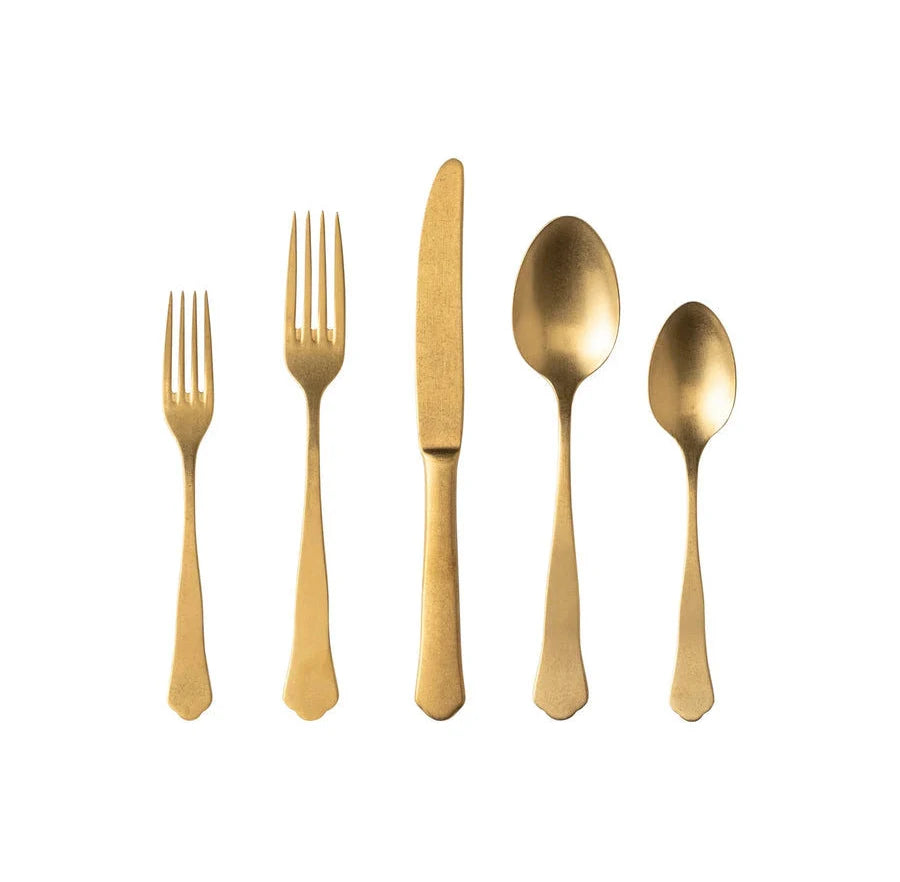 Five gold-colored Costa Nova "Prado - Gold" stainless steel utensils—a salad fork, dinner fork, knife, soup spoon, and teaspoon—are arranged in a row on a white background.