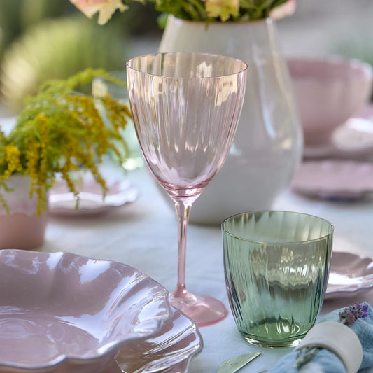 A close-up of an elegant table setting featuring the Casafina "Sensa" Rose-Magnolia wine glass, a green tumbler, pastel pink plates, and a white vase with yellow flowers on a white tablecloth.