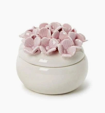 The Two's Company Hydrangea Trinket Box is a round white ceramic jar with a lid adorned by delicate, pale pink ceramic petals, capturing the hand-painted charm of hydrangeas and serving beautifully as a jewelry box.